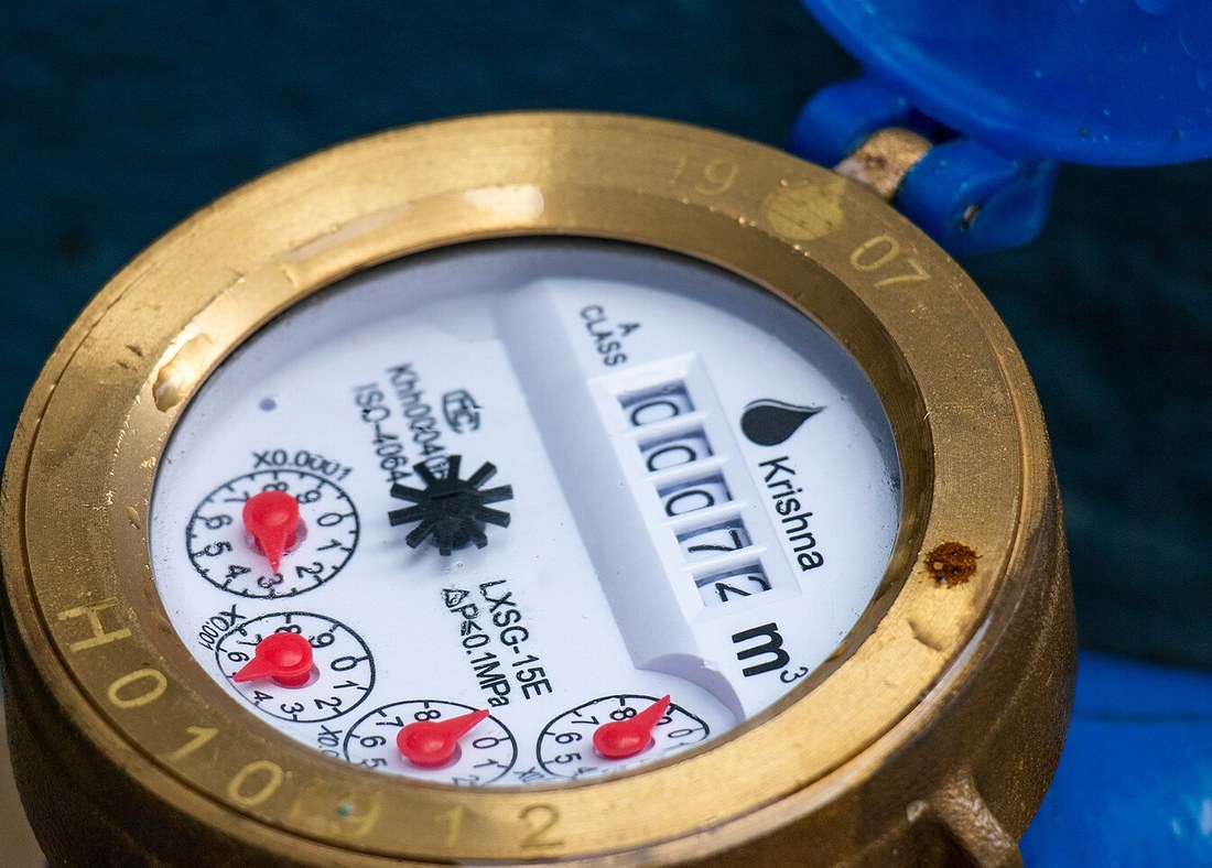 A brass Krishna water meter photographed at an angle. A vertical drum register shows the cubic metre count; four small dials with red pointers sit to its left.