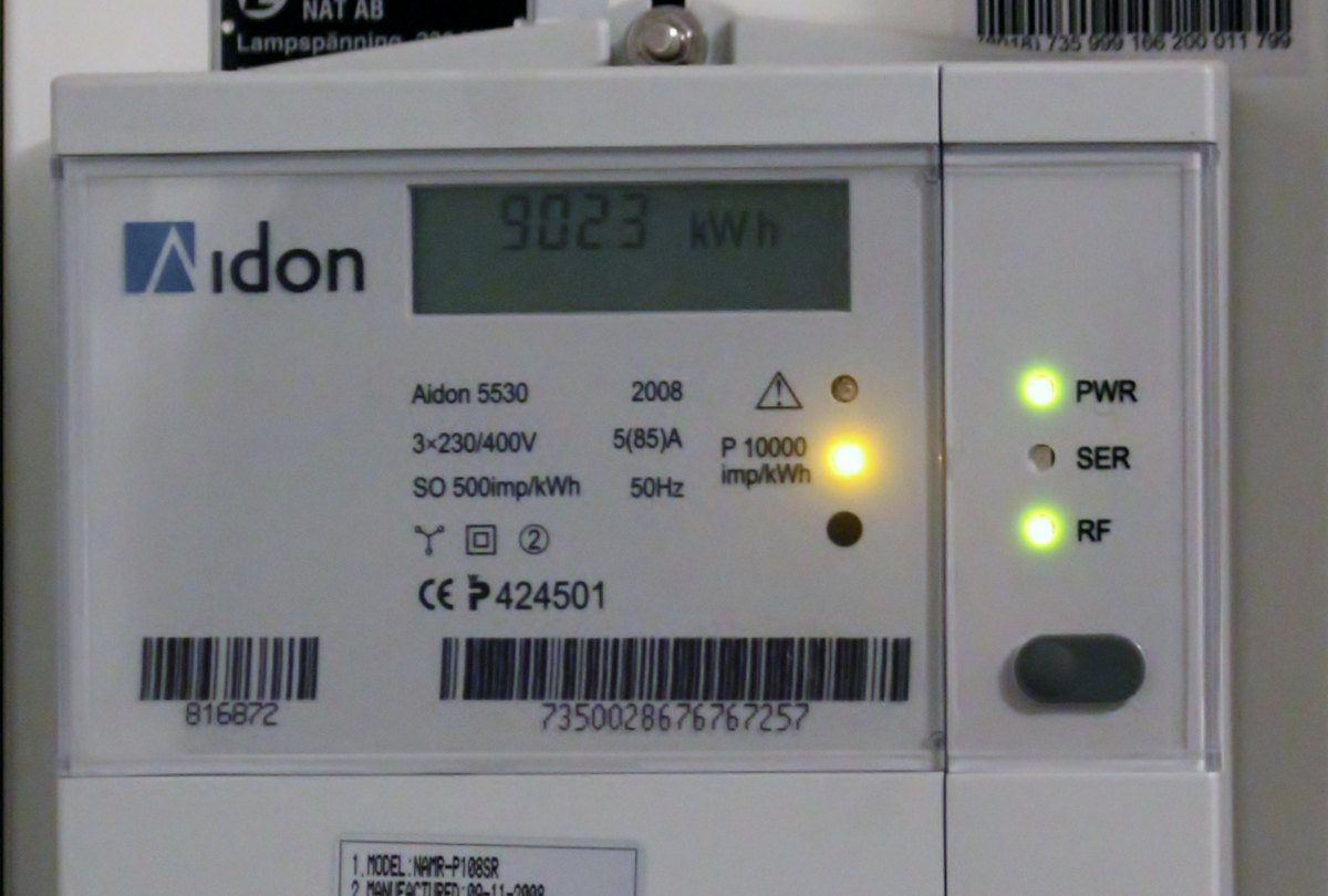 A wall-mounted Aidon electricity meter. The LCD reads 9023 kWh; below it are the model plate, a serial number and two barcodes.