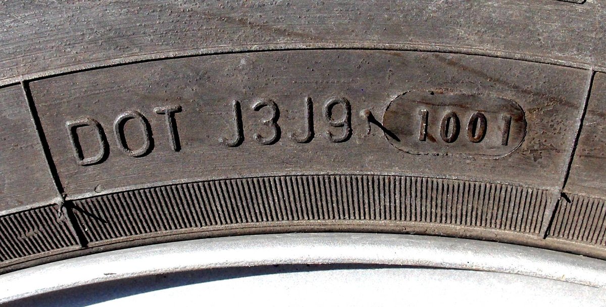 Close-up of a tyre sidewall stamped DOT J3 J9, a small tick, then the four-digit date code 1001 moulded inside its own oval.